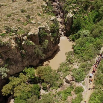 A still from The Age of Water. A birds-eye-view of a group of people hiking along a path that abuts a small river.