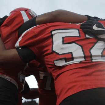 Still from Roll Red Roll. Close-up, an upward-angle shot of a few football players in red jerseys in a huddle.