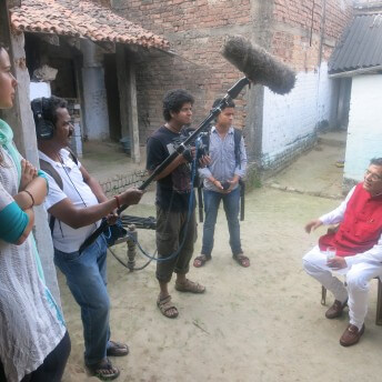 Still from Mister Toilet: The World's #2 Man. Three crew members stand outside filming a man sitting in a chair. There is a woman in the foreground leaning against a wall.