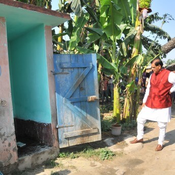 Still from Mister Toilet: The World's #2 Man. A few people stand around the outside of a small building. The door is open and a man is standing in front of it being filmed by a few crew members.