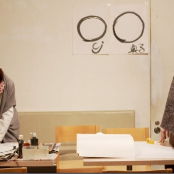 A still from The Departure. Two people are standing behind a desk with paper and ink sitting atop. Two hand-painted symbols hand on the wall in the background.