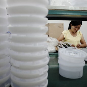Still from JOLIN: The Evolution of my Life. A woman is seen disassembling electronic parts. In the foreground is a close-up of a stack of white plastic containers.