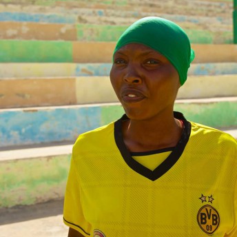 Still from Rajada Dalka/Nation's Hope. A woman stands in an exterior, in front of colorful, cement stadium-style seating steps. She is wearing a yellow jersey and a green headwrap.