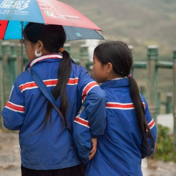 Still from Along the Line. Two young girls walk in the rain, one of them holds an umbrella, and both of them wear blue jackets.