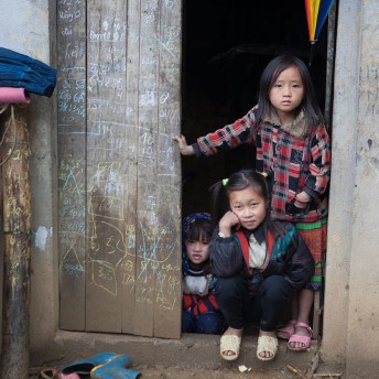 Still from Along the Line. Three small girls wearing sandals and warm jackets are standing behind an open wooden door. The door has chalk marks. The three girls are looking at the camera.