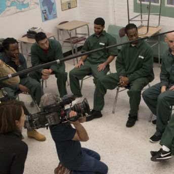 Production still of Lynn Novick's College Behind Bars: The Bard Prison Initiative. High angle shot of six men in prison uniforms sitting in a semi-circle inside of a classroom. A cameraman has a camera on his shoulder and films the men. Lynn the director is in the left corner looking at the shot. The audio pole is visible across the image.