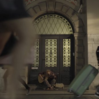 A person sitting cross-legged on a building's stoop looking down and playing an acoustic guitar, with three people in motion on the street in front of him