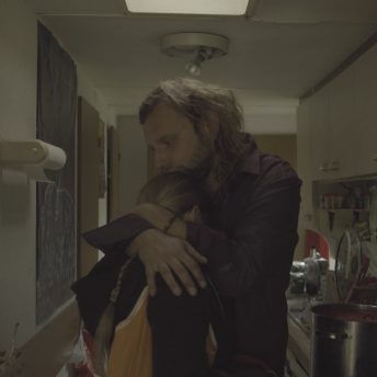 A blonde man and woman hugging in a small kitchen, with the woman's face hidden in the nook of the man's shoulder.