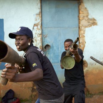Two persons point at the camera with metallic rusted look-alike guns. A slingshot is being held by a person out of the shot.