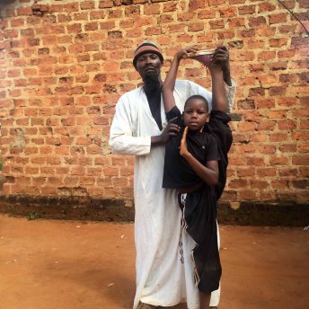 Still from Once Upon a Time in Uganda. A man stands behind and assists a young boy who is standing in a straddle position, standing on one foot with the other foot above his head. Behind them is a brick structure.