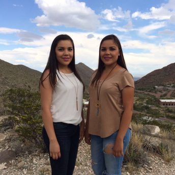 A still from The Guardian of Memory. Two people stand angled toward each other and look at the camera. They are standing in the middle of a plain, with large mountains off in the distance. The sun is low in the sky.