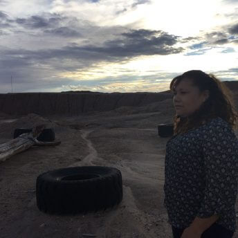 A still from The Guardian of Memory. A photo of a person in a dirt valley, looking off into the distance. There is a tire on the ground nearby.