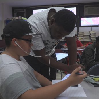 Still from Kids Can Spit. Two male high school students share wired earbuds. One student is sitting at a desk and holds a pen as he looks at a lined white sheet of paper. The other student is standing upright and leaning over the desk, also looking at the paper. Behind them, other students are sitting at desks.