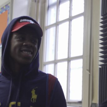 Still from Kids Can Spit. A male high school student dressed in a baseball hat and hoodie and wearing a backpack is smiling and talking to a man in glasses at the right-hand edge of the frame.