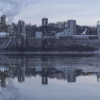 A still from The Devil We Know. A photo of a body of water. Across the water sits an industrial plant with smoke rising from columns.