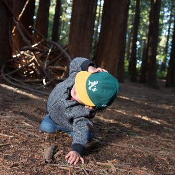Still from The F Word: A Foster-to-Adopt Story. A toddler dressed in jeans, grey jacket, and green-and-yellow baseball hat is kneeling on the forest floor, with one hand as support against the ground, the other hand against his head. The background is a light-speckled old-growth forest and directly behind him are a pile of branches.