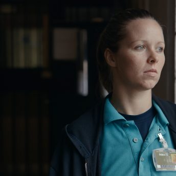 Still from Apart. Amanda awaits her judicial hearing, she is standing against a column.