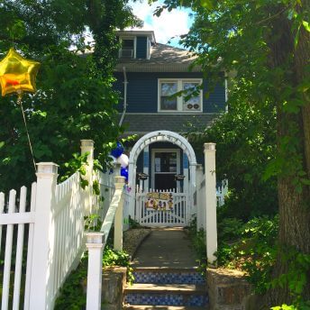 An Exterior shot of a blue house surrounded by green vegetation. There is a balloon in the shape of a golden star tied to the white fence in front of the house.