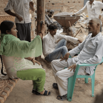 Still from Writing With Fire. A woman in green and white clothing sits and holds up a phone with a black case. A man sits opposite her and the camera, wearing all white, and sitting in a light blue chair. A few men and other materials are scattered behind them outside.