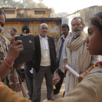 Still from Writing With Fire. Outside, a woman holds up a phone with a black phone case, while another woman stands in front of her. A few men stand behind them.