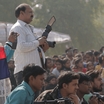Still from Writing With Fire. A crowd of people gathers outside. One man stands, wearing a light plaid shirt and holding a large camera. A few men sit in front of him, also holding cameras.