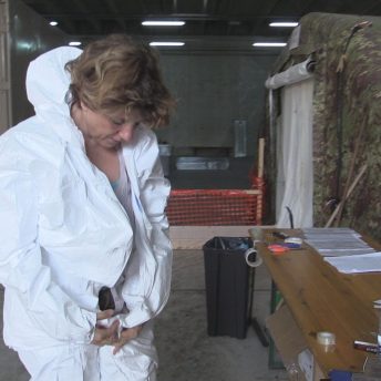 Still from Number 387. A woman zips up her medical hazard suit. She is standing in front of a table and military tent structure.