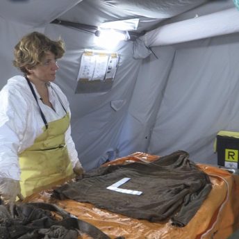 Still from Number 387. Two women in a medical tent stand at a table across from each other wearing PPE. One woman has a hand raised. There is an item of clothing on the table with an "L" ruler on top of it.
