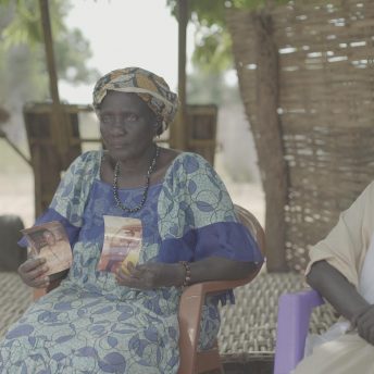 Still from Number 387. A man and woman are sitting outside in chairs. The woman is holding a picture in each hand.