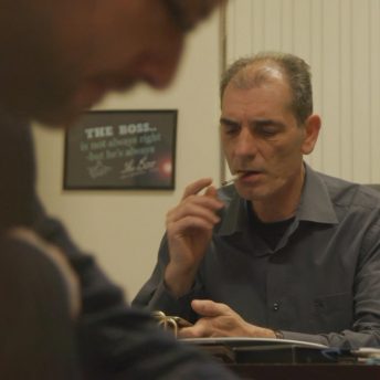 Still from Number 387. A man is sitting at a desk smoking a cigarette and holding a cell phone. There is an out of focus person in the foreground.