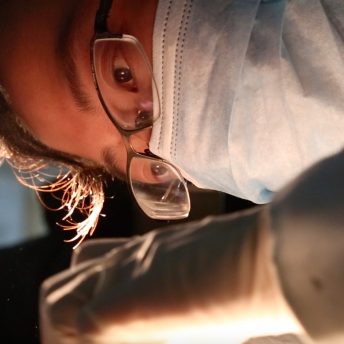 Close-up of a person wearing a surgical mask looking at their hand holding something out of view with a light shining on it