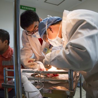Two medical staff in lab coats hover over a brightly lit tray of bloody gauze and a man in an orange jacket sits nearby
