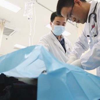 Two medical staff work on or over a body lying on a hospital bed covered by a blue sheet