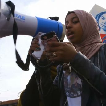 Still from Accept the Call. Low angle shot of a young girl speaking through a megaphone while reading from a cellphone. She is wearing a hijab, a t-shirt, and a jacket.