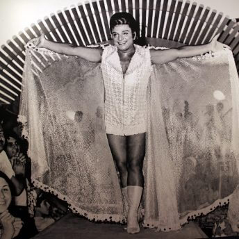 Still from A Prince from Outer Space: Zeki Muren. A man wearing a jumper and tall boots stands on a stage with a fabric cape draping from his hands. There is a crowd of people surrounding him.