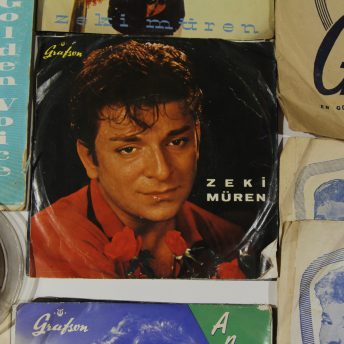 Still from A Prince from Outer Space: Zeki Muren. The record album of Zeki Muren is sitting on a table with other publications and media. On the record is an image of Zeki Muren with some red roses.