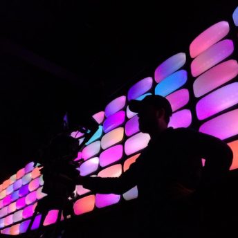 A man wearing a cap is standing in front of a wall full of pink and purple lights, next to him is a camera on a tripod.