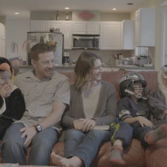 Still from Made in Boise. A man, woman, and three children in costumes sit on a couch, smiling. There is an out of focus kitchen in the background.
