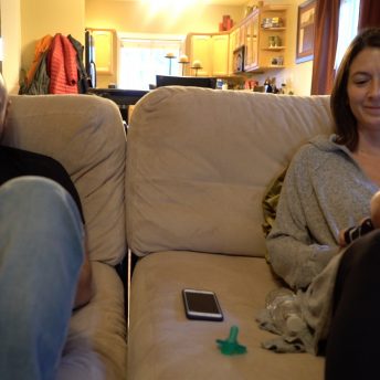 Still from Made in Boise. A man and a woman sit on a couch, each holding an infant, smiling. The background is a well lit, out of focus dining room and kitchen.