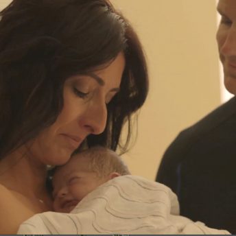 Still from Made in Boise. A woman is holding an infant, covered by a blanket. A man stands on her left, smiling and looking at the infant in her arms.