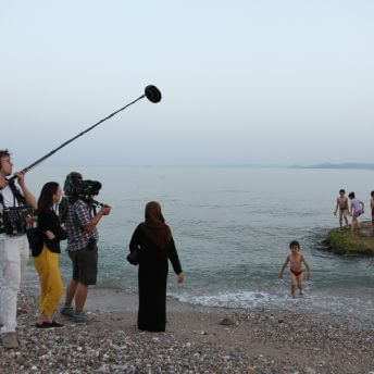 Production still from As Simple As Water. Director Megan Mylan, and her cameraman and audio person, follow a family playing on the ocean shore. The mother waits while the children play in the water.