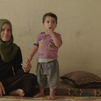 A mother wearing a hijab sits on a carpeted floor and holds the hand of her toddler son. Both of them look away from the camera.