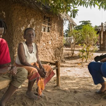 Still from The Letter. Two people sit in front of a house constructed of clay walls. It is sunny outside and the sky is blue.
