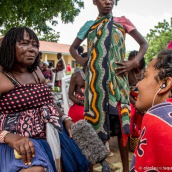 Still from The Letter. A group of people outside in the yard in front of a house. They sit in small groups. All are wearing different versions of colorful patterned cloth. Three people are situated in the foreground of the image. Two sit on either side of the frame, facing each other. The third stands between them, hands on hips.