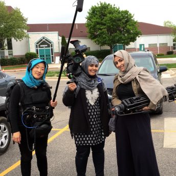 Production still of An Act of Worship. Three women crew members are standing in a parking lot, wearing hijabs, and holding film equipment.