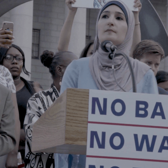 Still from An Act of Worship. A woman wearing a hijab is speaking in front of a podium, the podium has a poster that says "no ban, no wall." Behind her, there is a group of people.