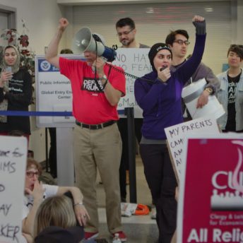 Still from An Act of Worship. People participating in a protest against the Muslim ban, the hold posters.
