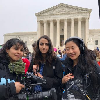 Production still of An Act of Worship. Three women dressed in black of the film's production crew are standing in front of the United States Supreme Court, director Nausheen Dadabhoy, to the left, is holding a camera.
