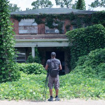 A man standing in front of a house surrounded by green vegetation, with a camera tripod in front of him.