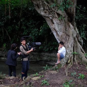 Production still from Silent Beauty. Two persons, one with a camera and the other with a headshot work recording a young boy leaning on a big tree. They are surrounded by nature.