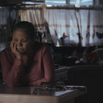 A woman is leaning on a kitchen counter. She holds her face with her left hand and looks away from the camera.